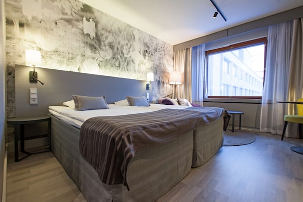 Una habitación de hotel moderna con una cama grande, mesitas de noche, un ventanal con cortinas y un mural con vista a la ciudad.