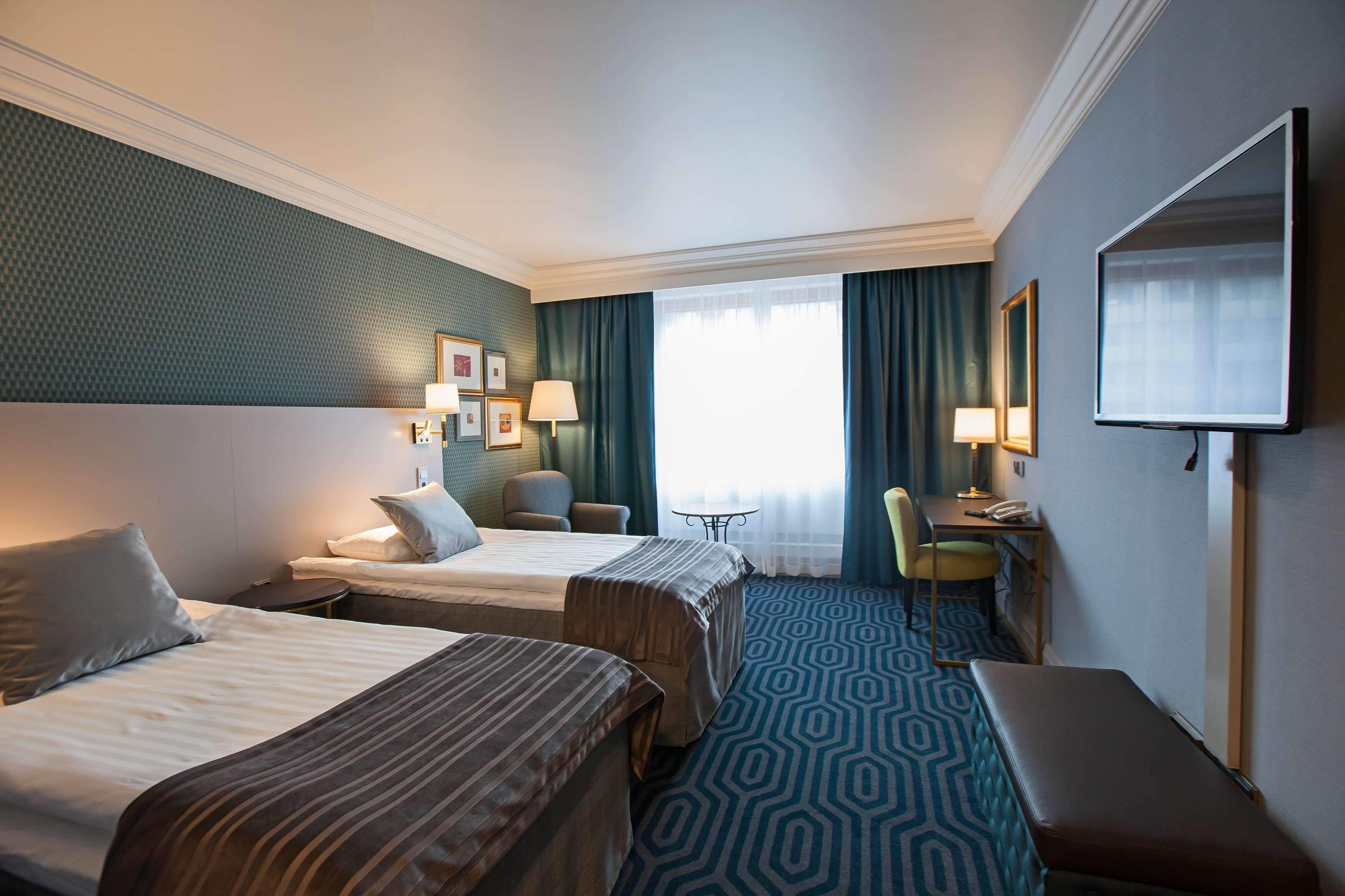 A hotel room with two beds, a desk, a chair, a bench, and a TV.