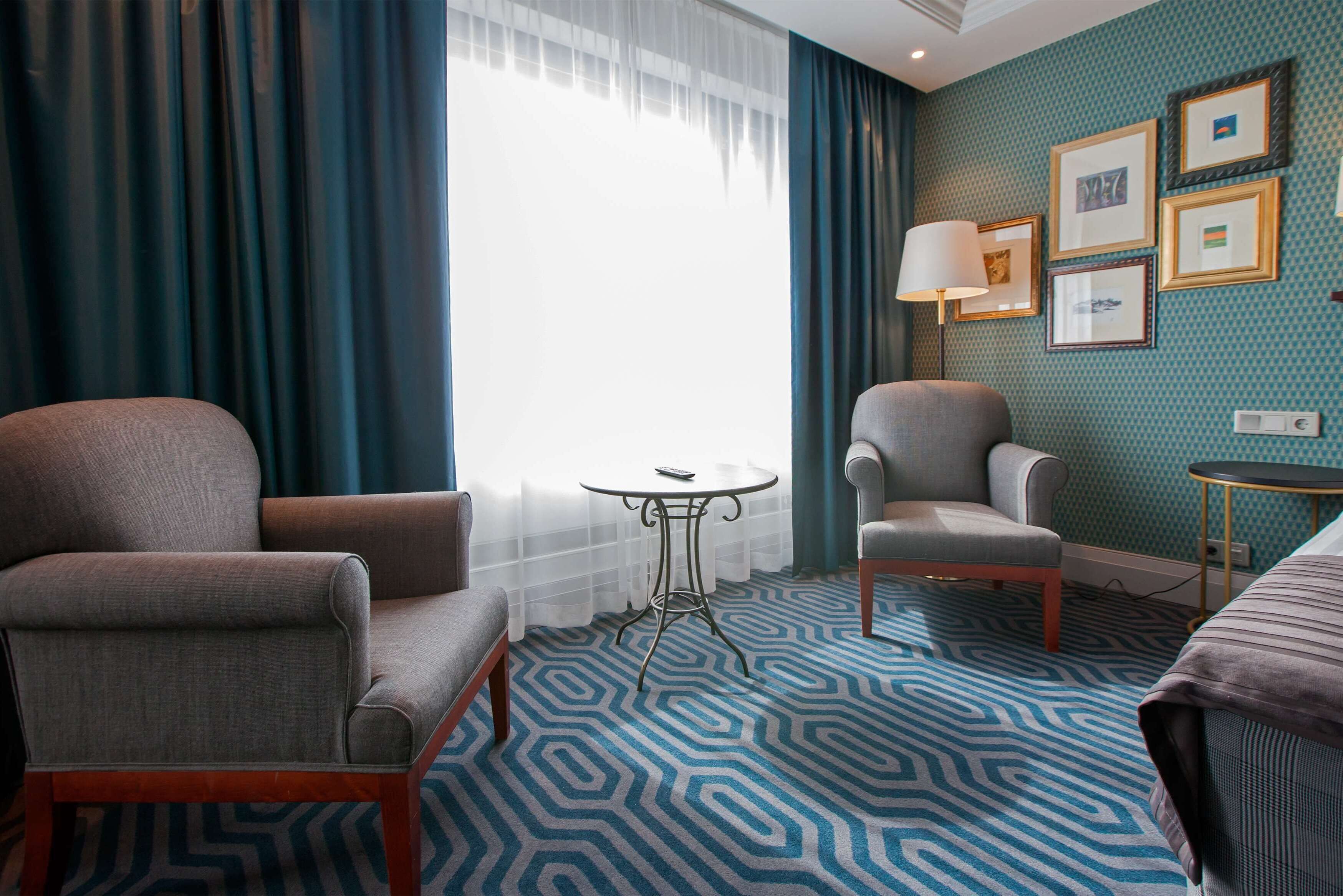 Ein Hotelzimmer mit Teppichmuster, zwei Sesseln, einem kleinen runden Tisch, einer Lampe und einem Bett.