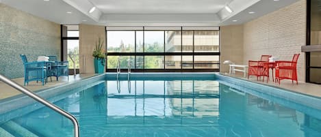 An indoor swimming pool with clear blue water, surrounded by blue and red chairs, and large windows offering a view of greenery outside.