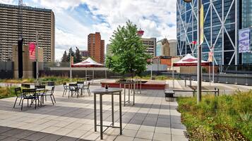 An outdoor seating area with tables and chairs, surrounded by greenery and modern buildings in the background.