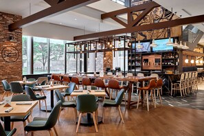 Um restaurante moderno com mesas e cadeiras de madeira, um espaço de bar e grandes janelas com vista para o exterior.
