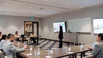 A conference room with a presentation screen, a whiteboard, and attendees seated at a long table with water bottles and notepads.