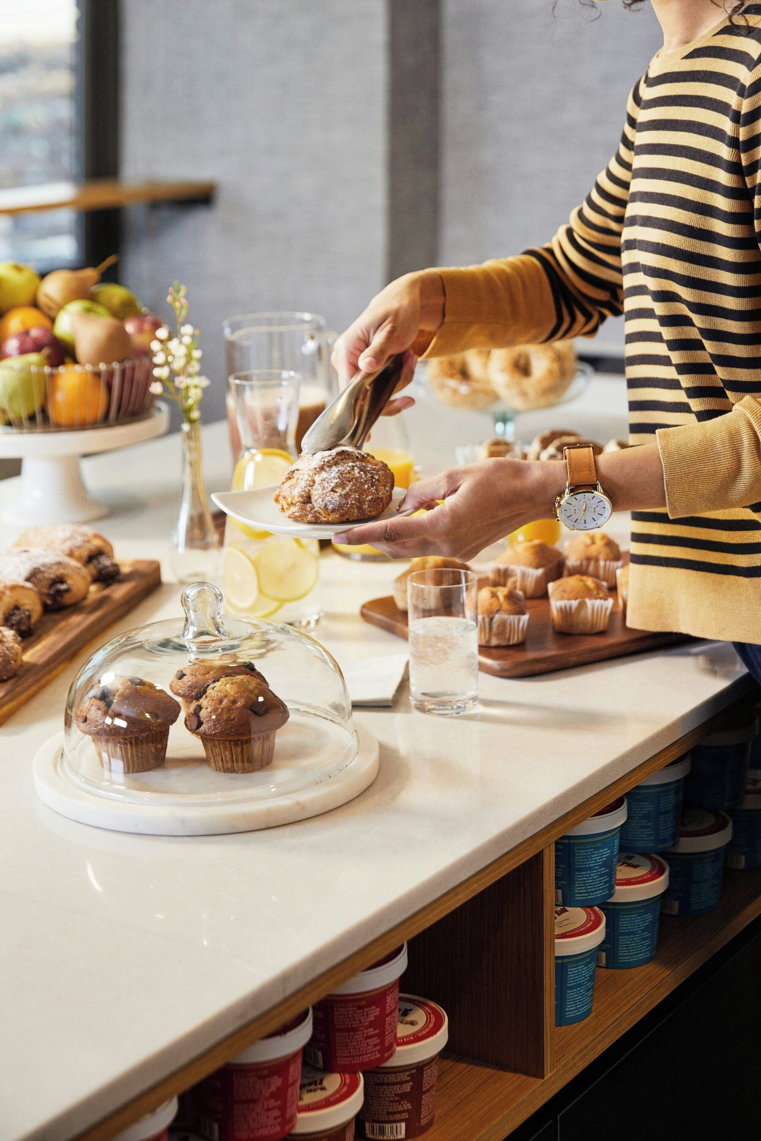Una persona sirviendo muffins en un mostrador con una variedad de productos horneados y bebidas.