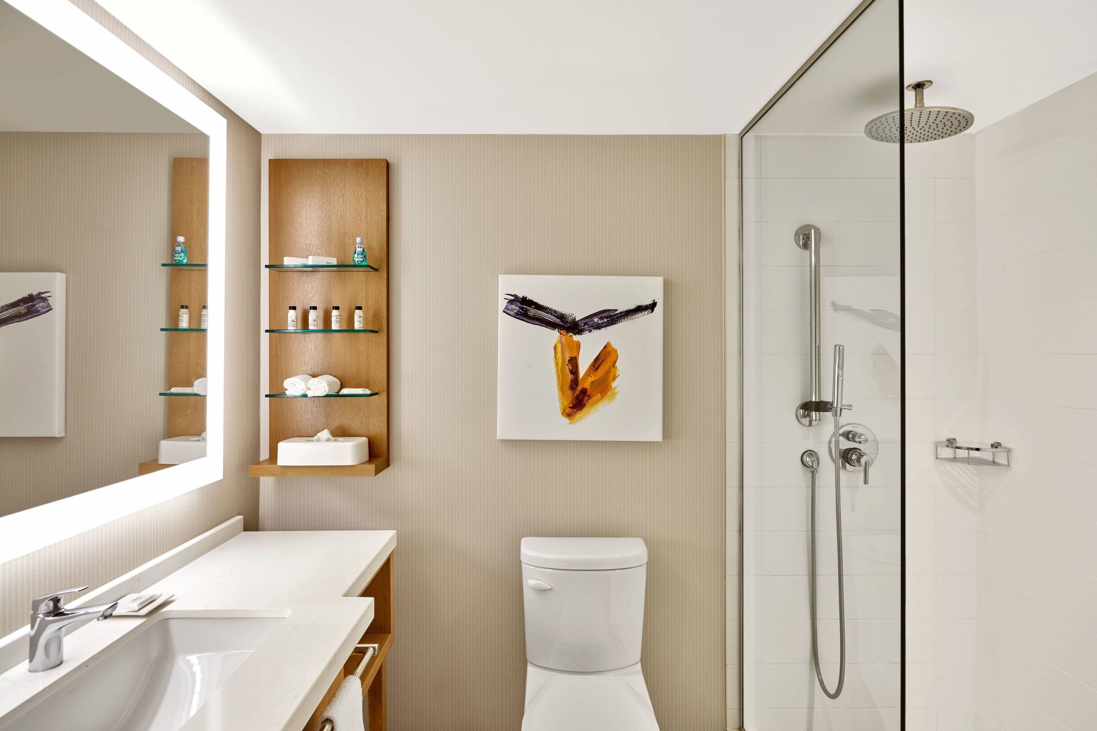 Un baño moderno con lavamanos, espejo y retrete blancos. Cuenta con una ducha con cabezal de ducha de mano y mampara de vidrio. Una repisa de madera con artículos de aseo está montada en la pared.