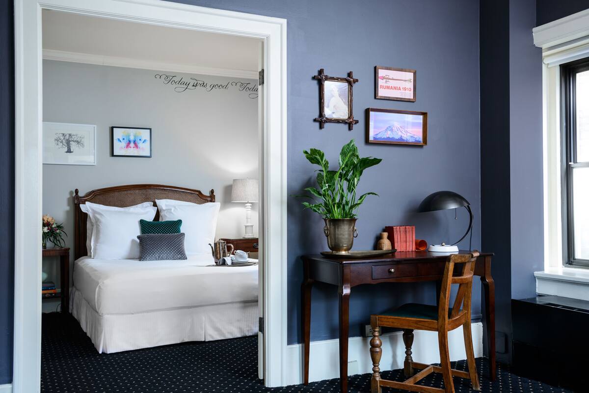 Un dormitorio con cama, mesita de noche, lámpara y escritorio con silla. En la pared hay cuadros enmarcados y una planta en maceta sobre el escritorio.