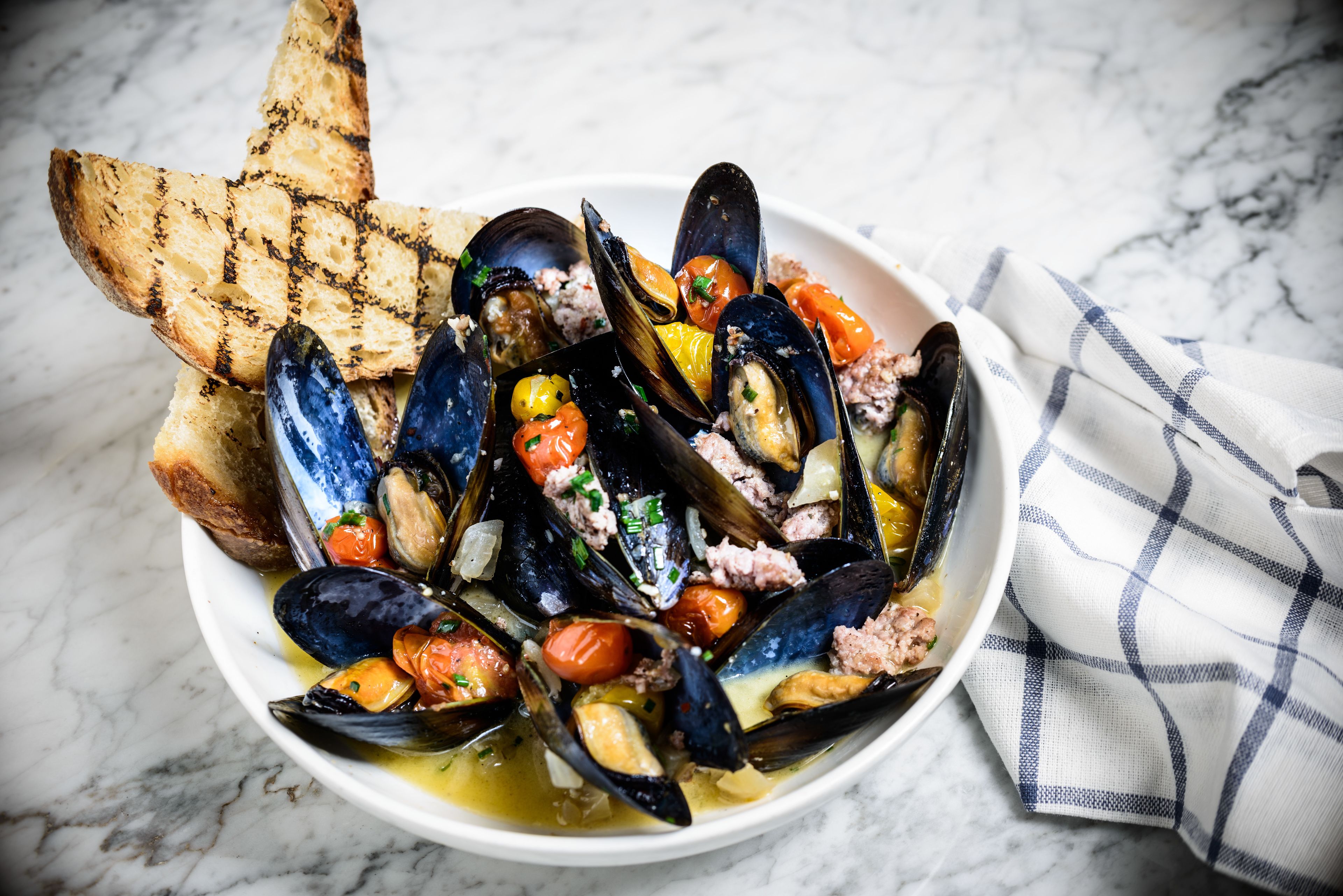 Un bol de mejillones con tomates y pan sobre una superficie de mármol.