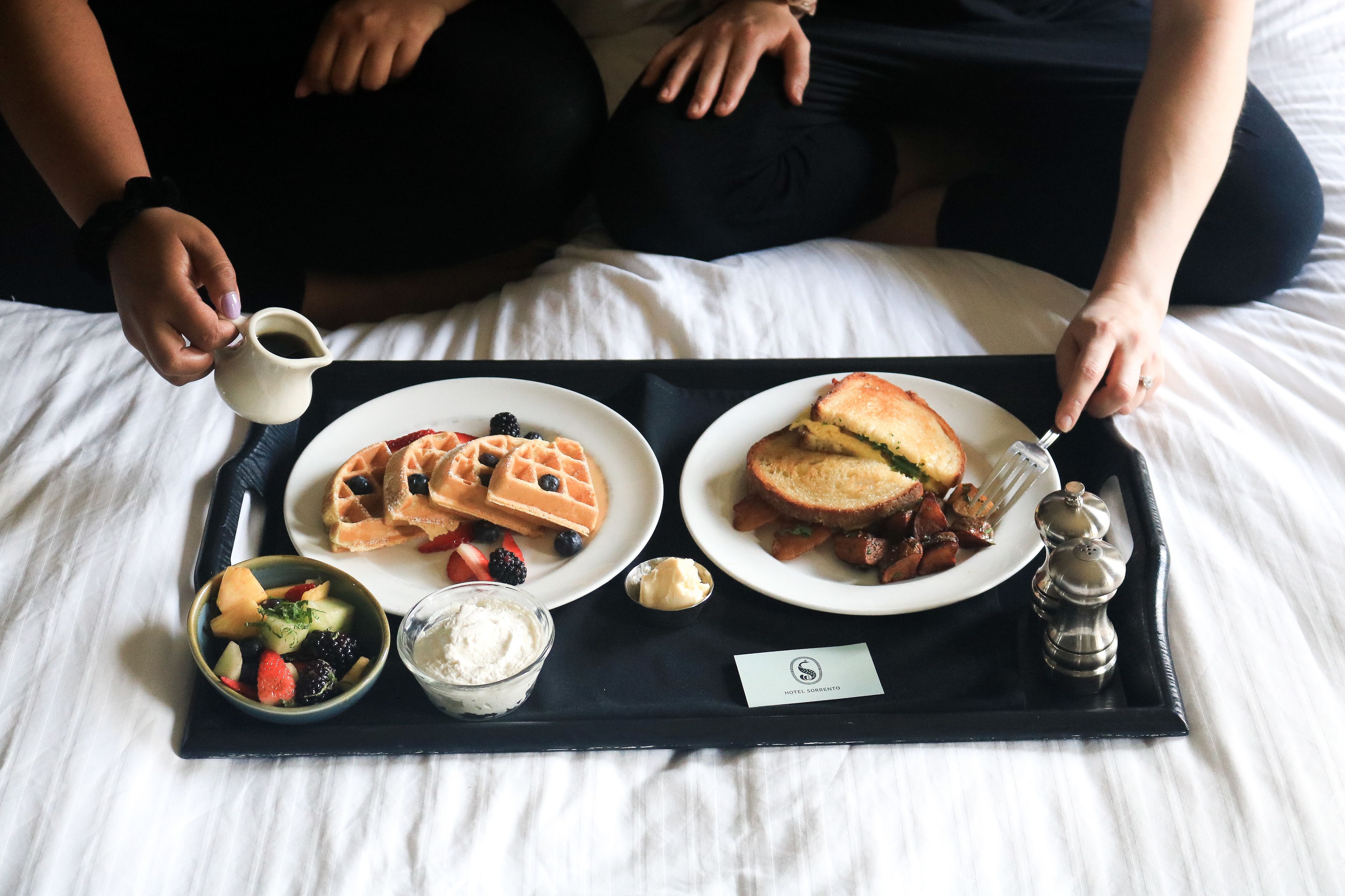 Ein Tablett mit Waffeln, einem Sandwich, Obst und Beilagen auf einem Bett.