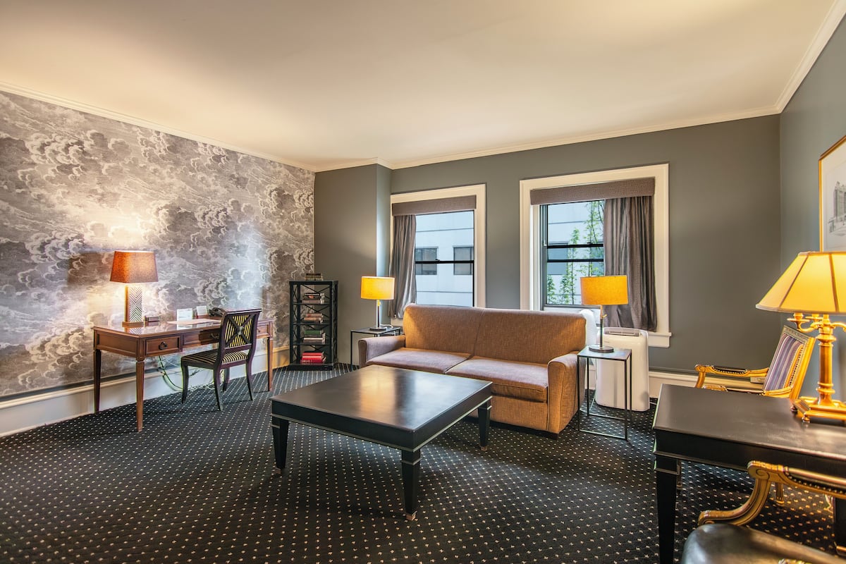 Habitación de hotel con sofá, una mesita de centro, escritorio y dos lámparas. Cuenta con dos ventanas con cortinas, una estantería y una alfombra estampada.