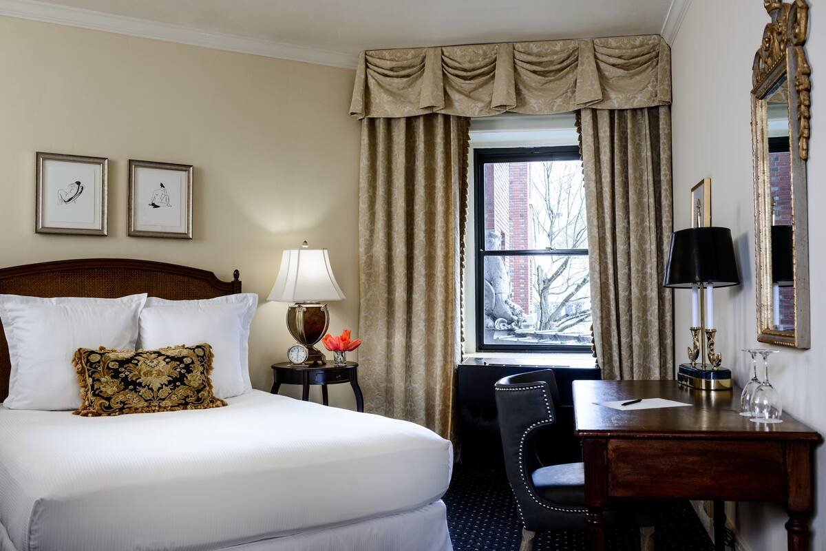 Habitación de hotel con cama, escritorio, silla, lámpara, reloj y ventana con cortinas.