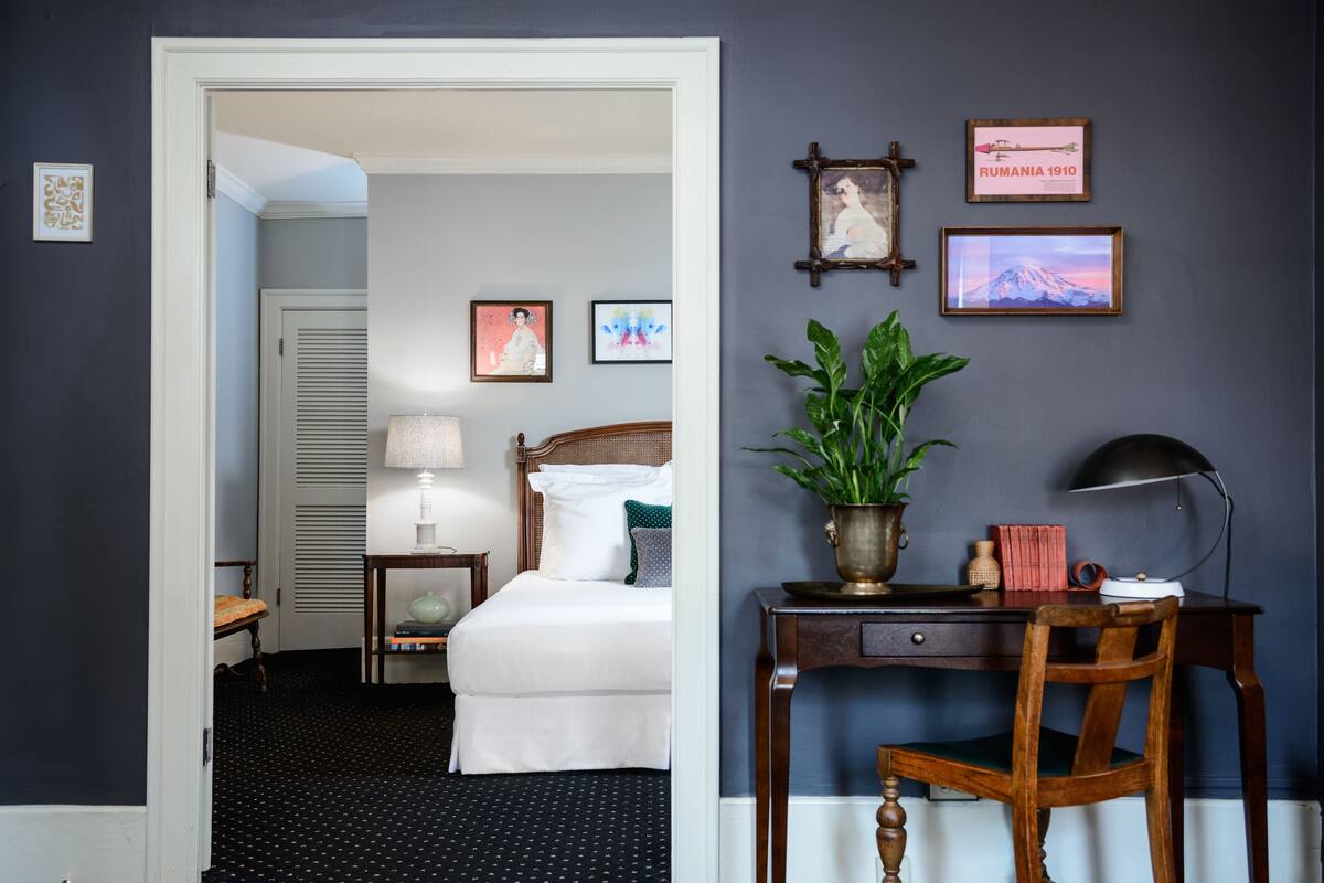 Habitación de hotel con cama, escritorio con lámpara, silla y planta en maceta. Se observan cuadros en la pared y una puerta que da a otra habitación.
