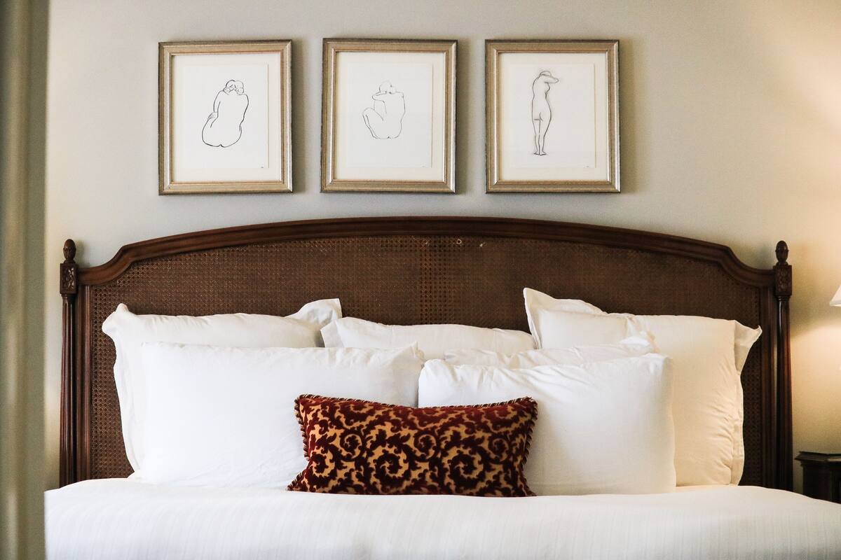 Una cama bien hecha con sábanas blancas y una única almohada estampada, una obra de arte enmarcada en la pared encima y un cabecero de madera.