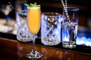 A bar setting with a tall glass of a yellow beverage garnished with a mint leaf, surrounded by ice-filled glasses.