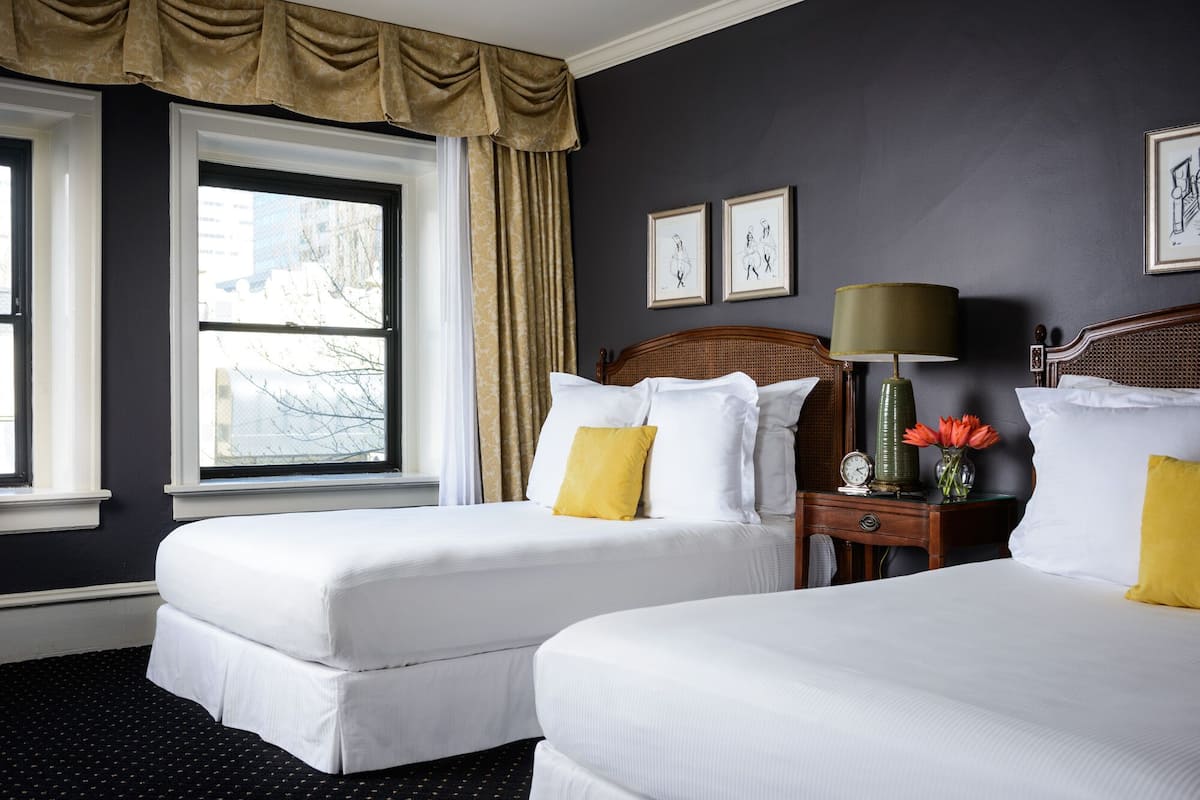 Habitación de hotel con dos camas, una mesita de noche, una lámpara y un ventanal con cortinas.