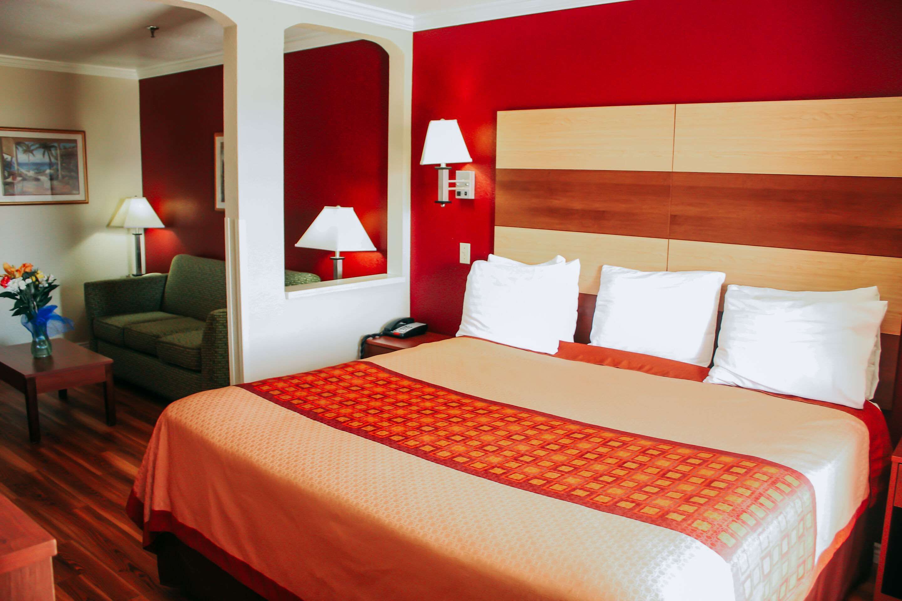 A hotel room with a large bed, a green armchair, a nightstand with a lamp, and a wall-mounted mirror.