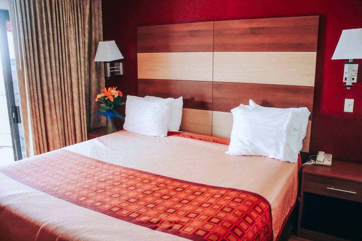 Ein Hotelzimmer mit einem großen Bett, zwei weißen Kissen, einer roten und orangefarbenen Tagesdecke, einem Holzkopfschild, einem Nachttisch mit einer Lampe und einer Vase mit Blumen.