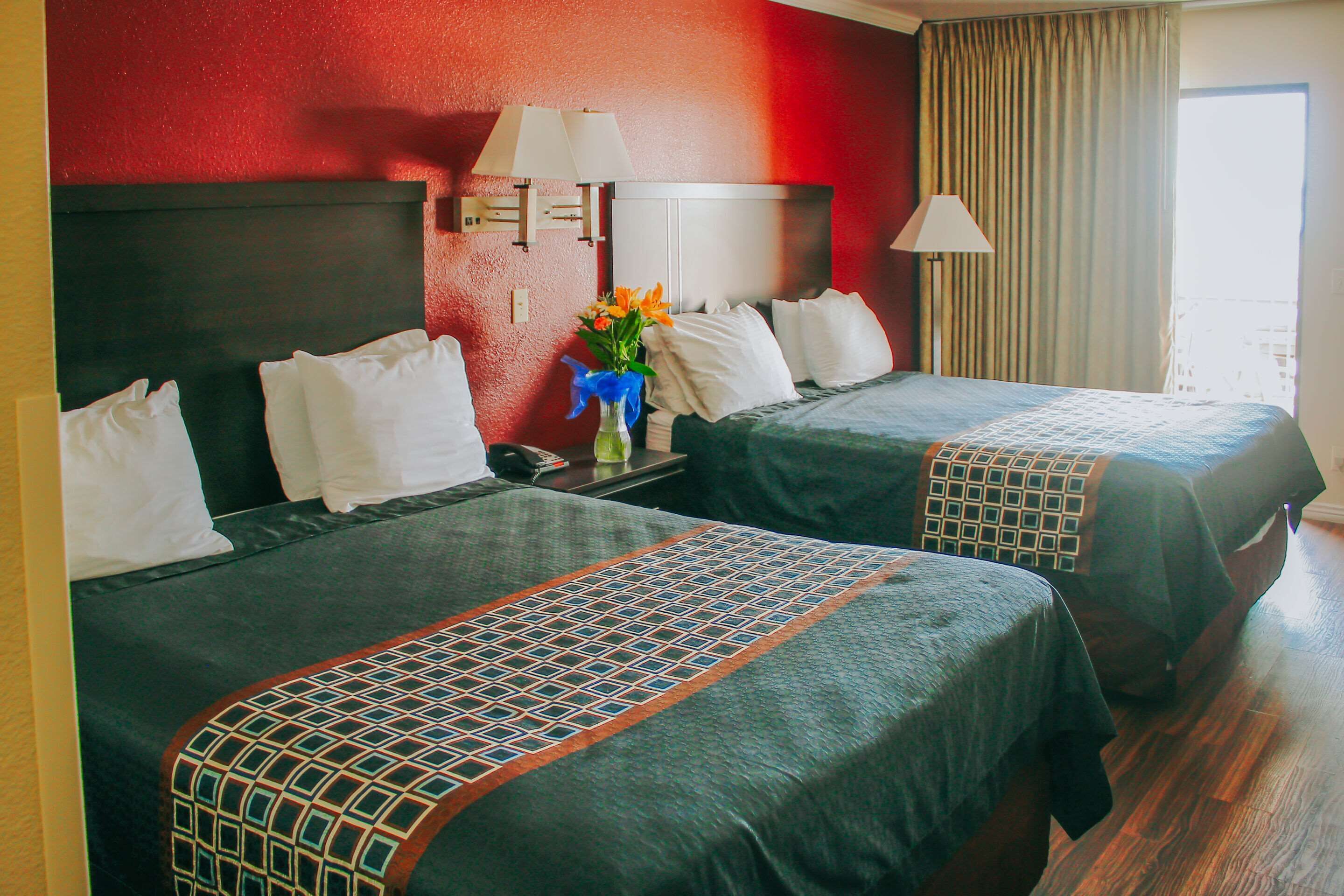 A hotel room with two beds, each with a patterned bedspread and white pillows. There is a nightstand with a lamp and a vase with flowers. The room has a red accent wall and a window with curtains.