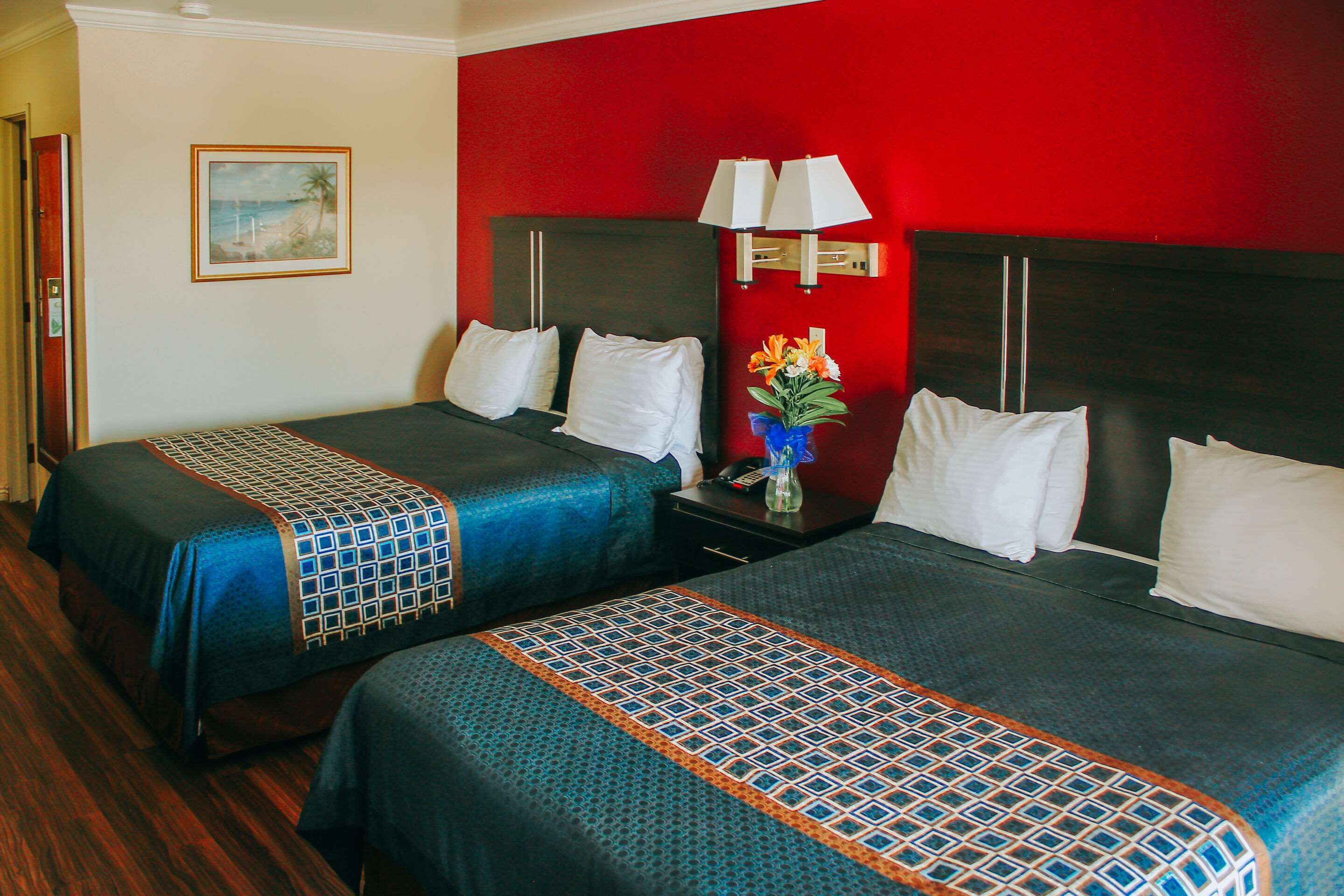 Habitación de hotel con dos camas, una mesita de noche con lámpara, un cuadro en la pared y una pared de acento en rojo.