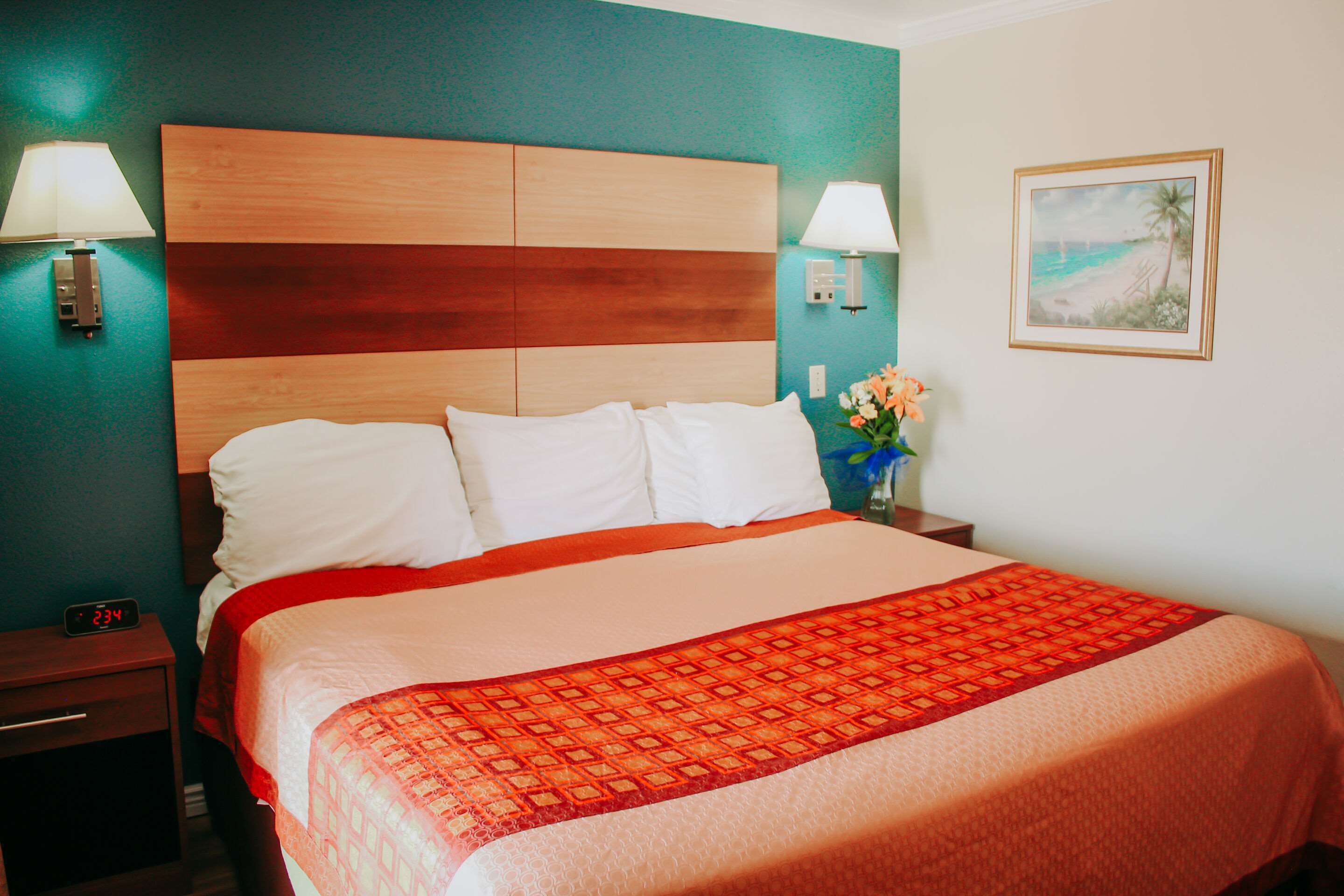 A hotel room with a large bed, two bedside tables, a nightstand, a wall clock, a framed picture, and a vase with flowers.