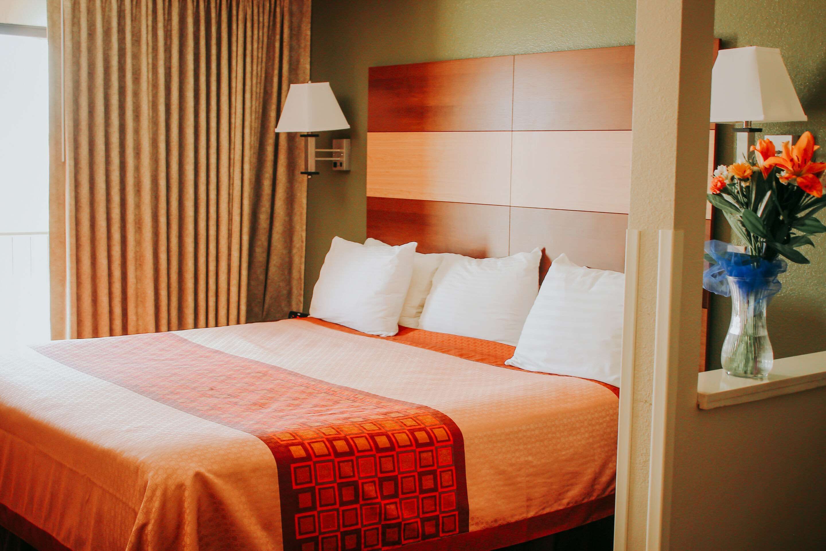 A hotel room with a large bed, a headboard, two wall-mounted lamps, a vase with flowers, and a window with curtains.