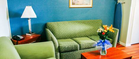 Quarto com sofá verde, mesa de centro de madeira com um vaso de flores, luminária e um quadro na parede.