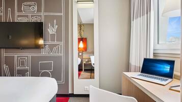 Habitación de hotel con una cama, un escritorio con una computadora portátil, un televisor y vistas a la ciudad a través de la ventana.