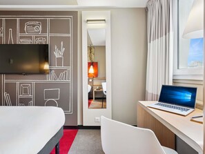Hotellihuone, jossa on sänky, työpöytä kannettavalla tietokoneella, televisio ja ikkunasta avautuva näkymä kaupunkiin.