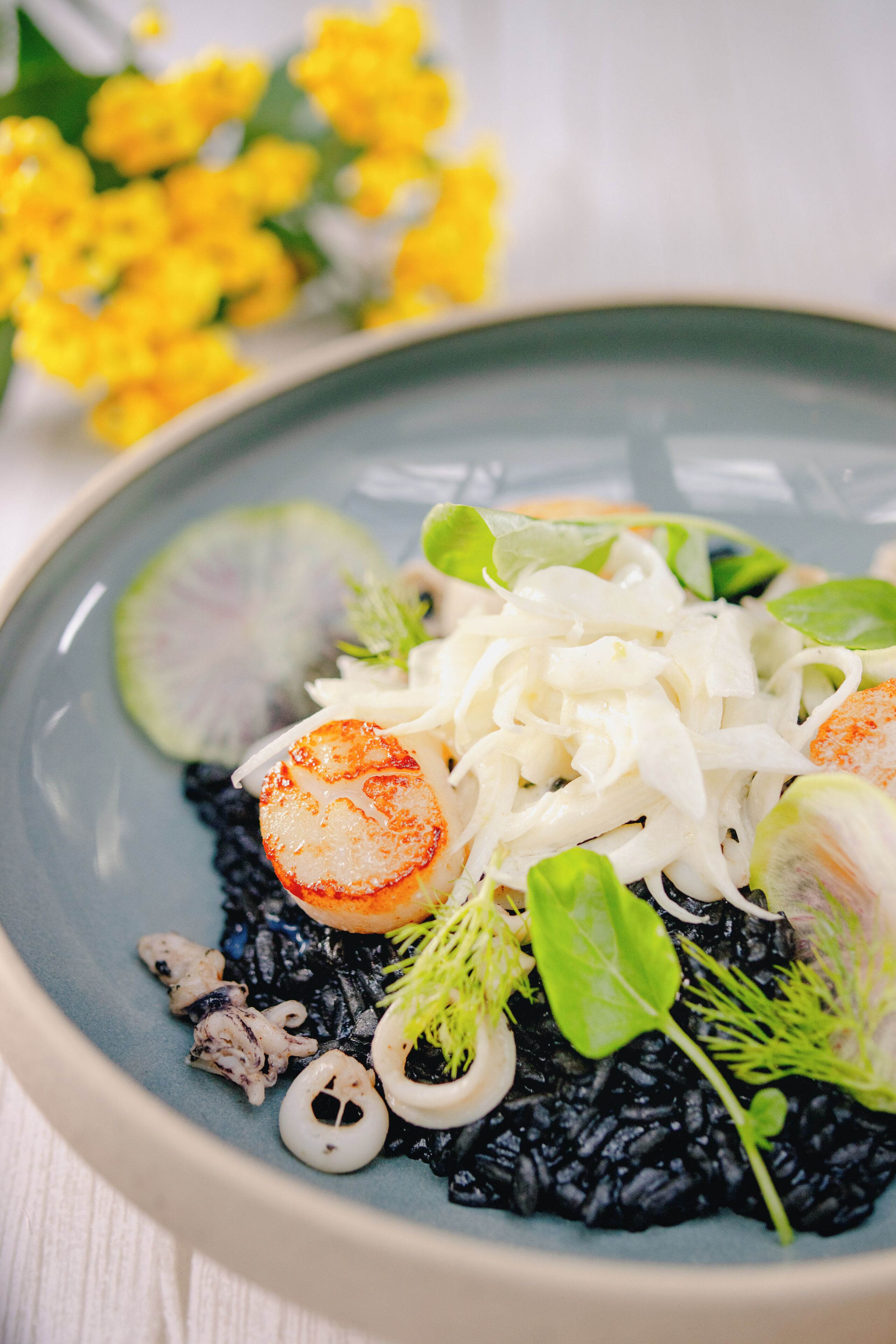 Un plato de risotto negro con vieiras, verduras de hoja y una rodaja de pepino.
