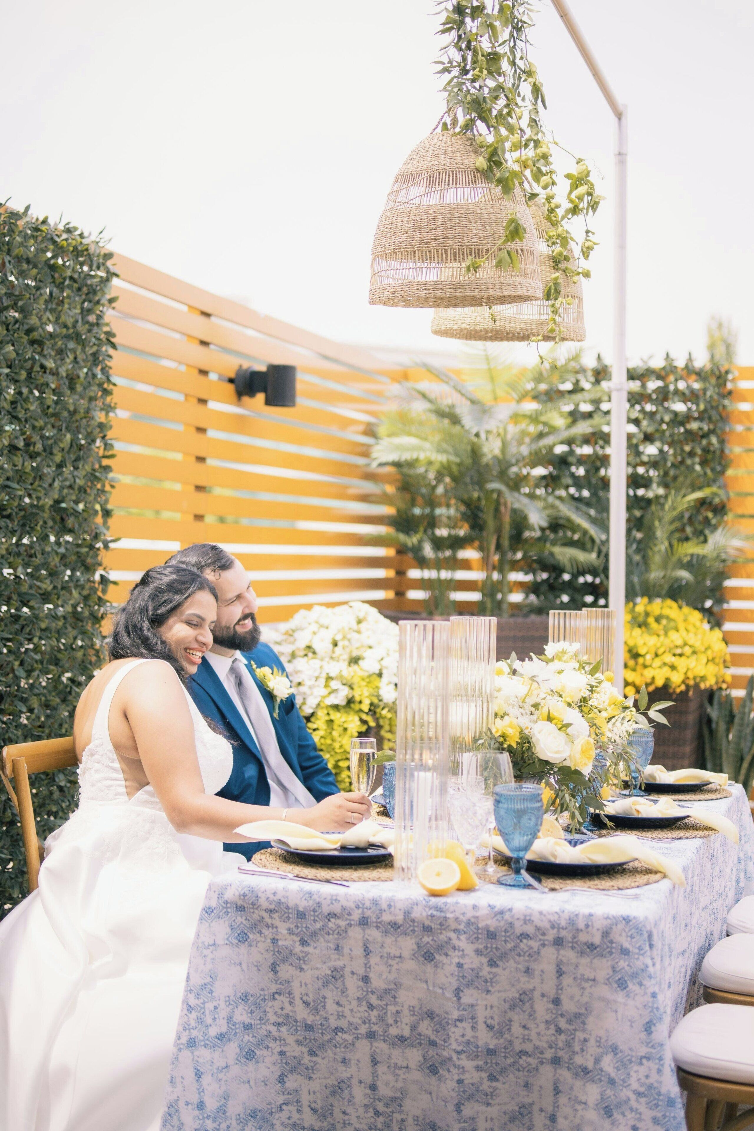Een stel zit aan een tafel met een blauw geruit tafelkleed, omringd door groen en gele bloemen.