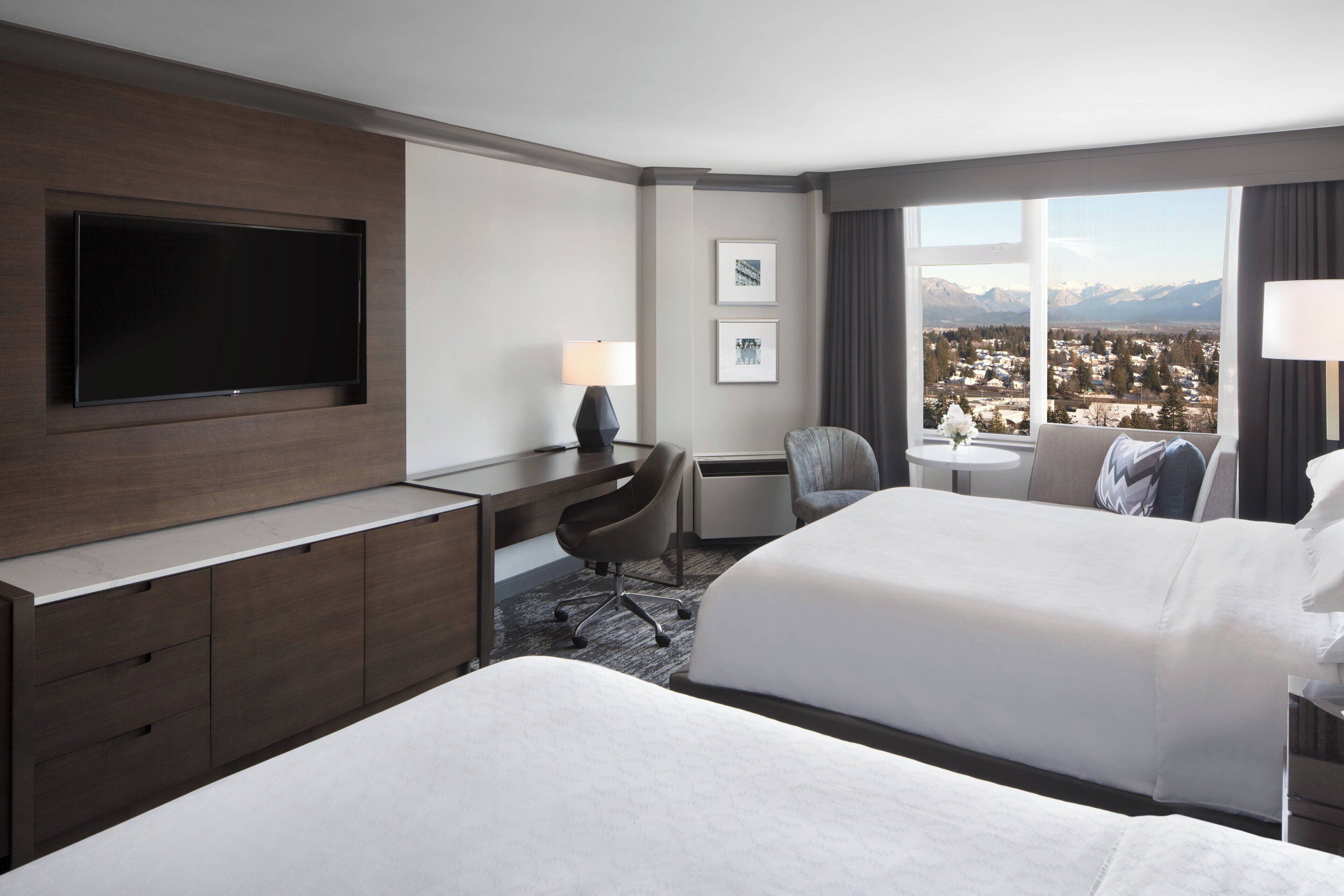 Habitación de hotel con dos camas, un escritorio con televisor de pantalla plana, una silla, una mesita y vistas a un paisaje nevado a través de amplios ventanales.