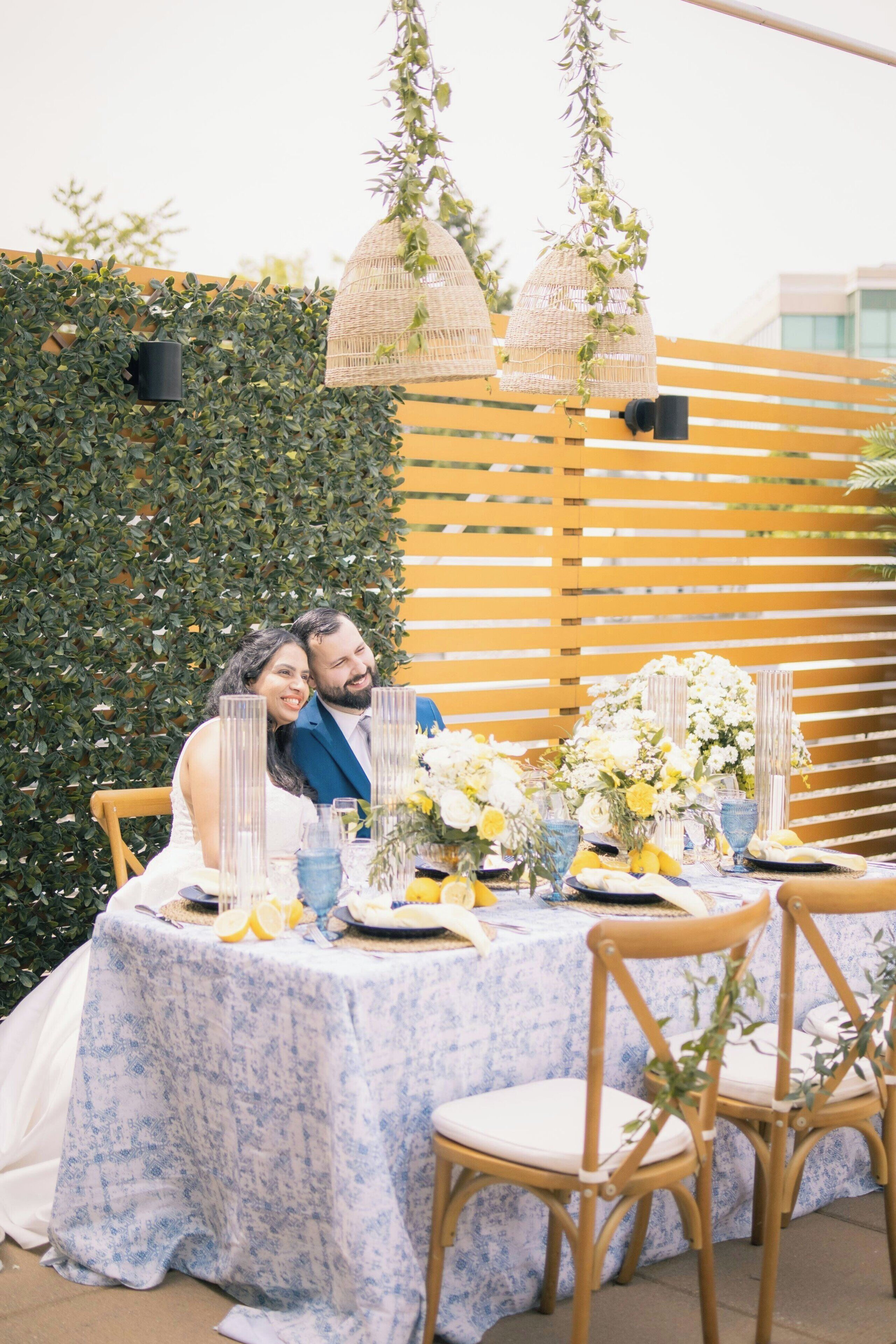 Una pareja sentada en una mesa al aire libre con mantel floral azul, rodeada de follaje y listones de madera amarillentos.