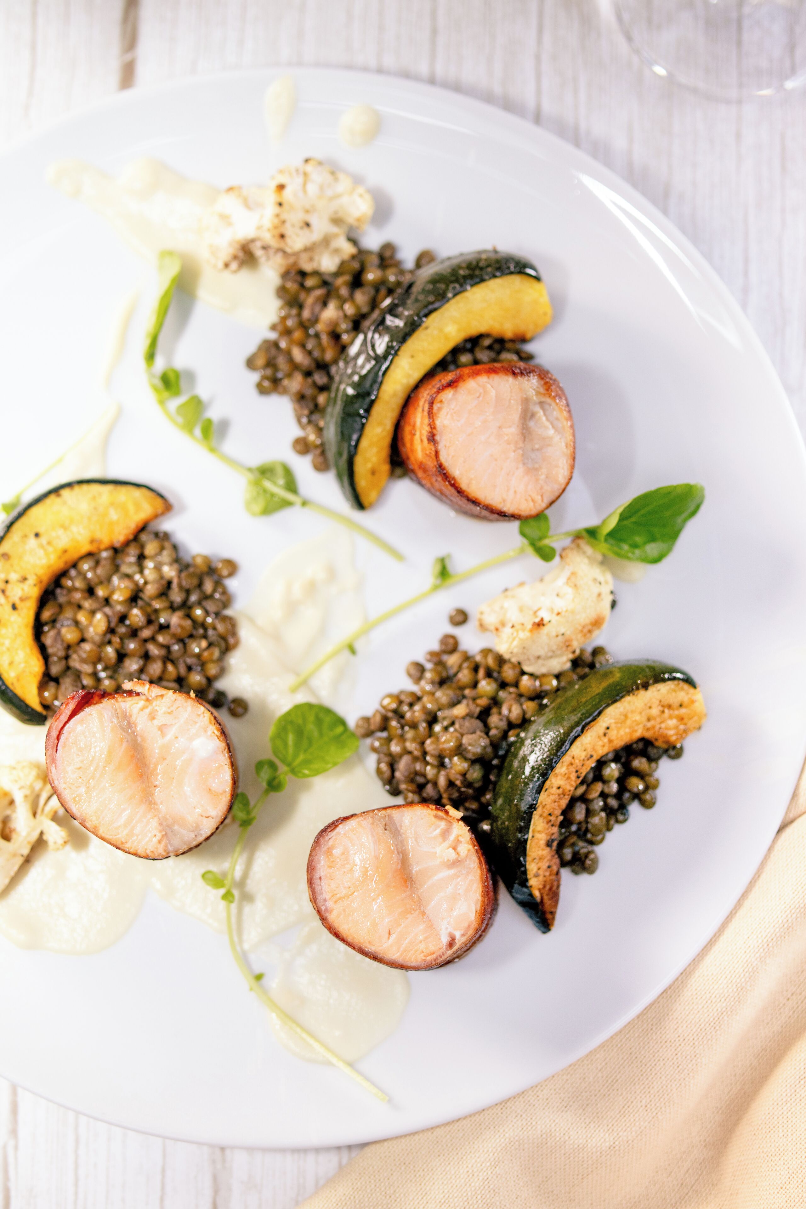 Un plato blanco con diversos alimentos, incluyendo verduras y un trozo de carne, posiblemente salmón, adornado con hojas verdes.