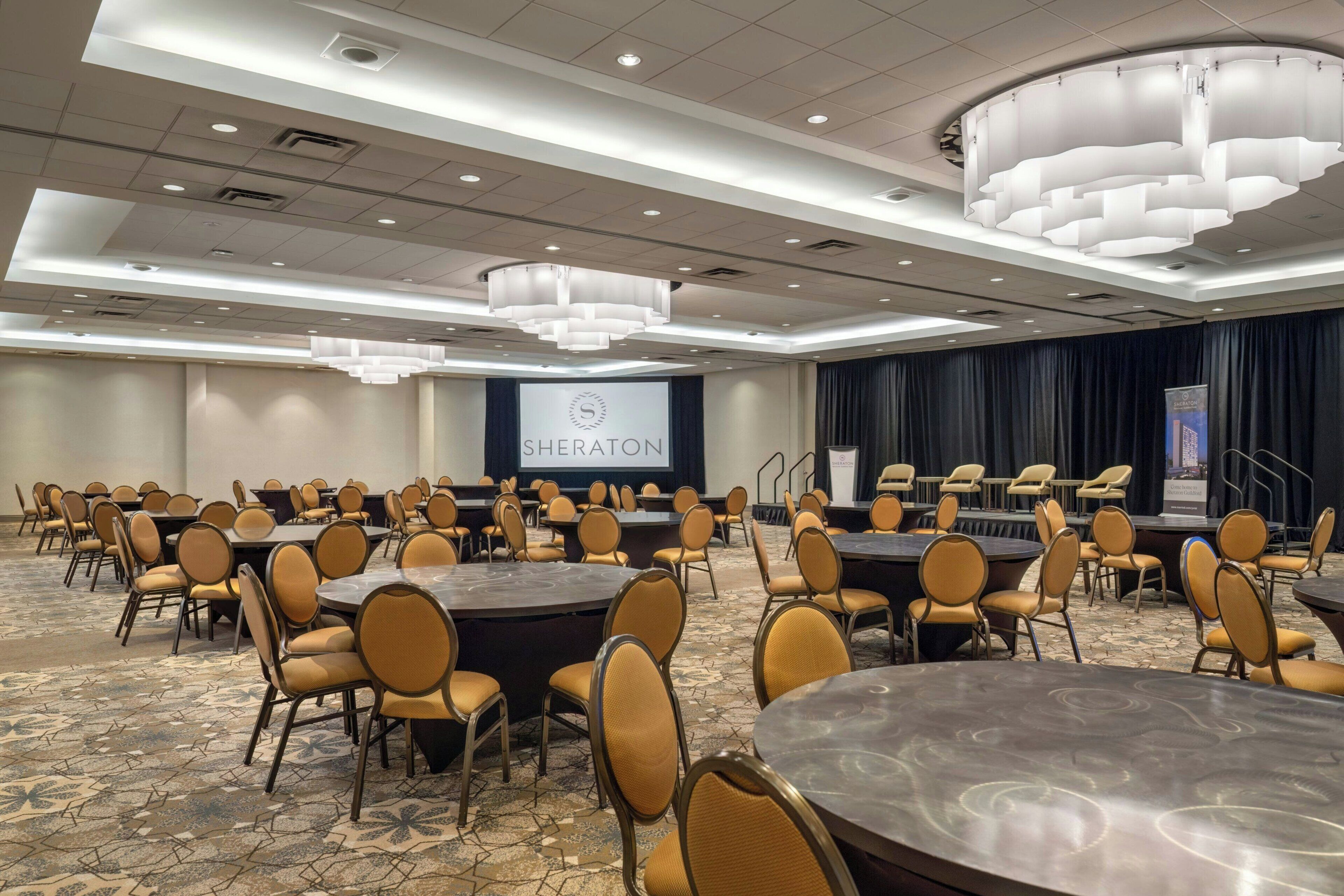 Une salle de conférence avec des tables rondes et des chaises, un grand écran affichant le mot « Sheraton », et une scène avec des rideaux.
