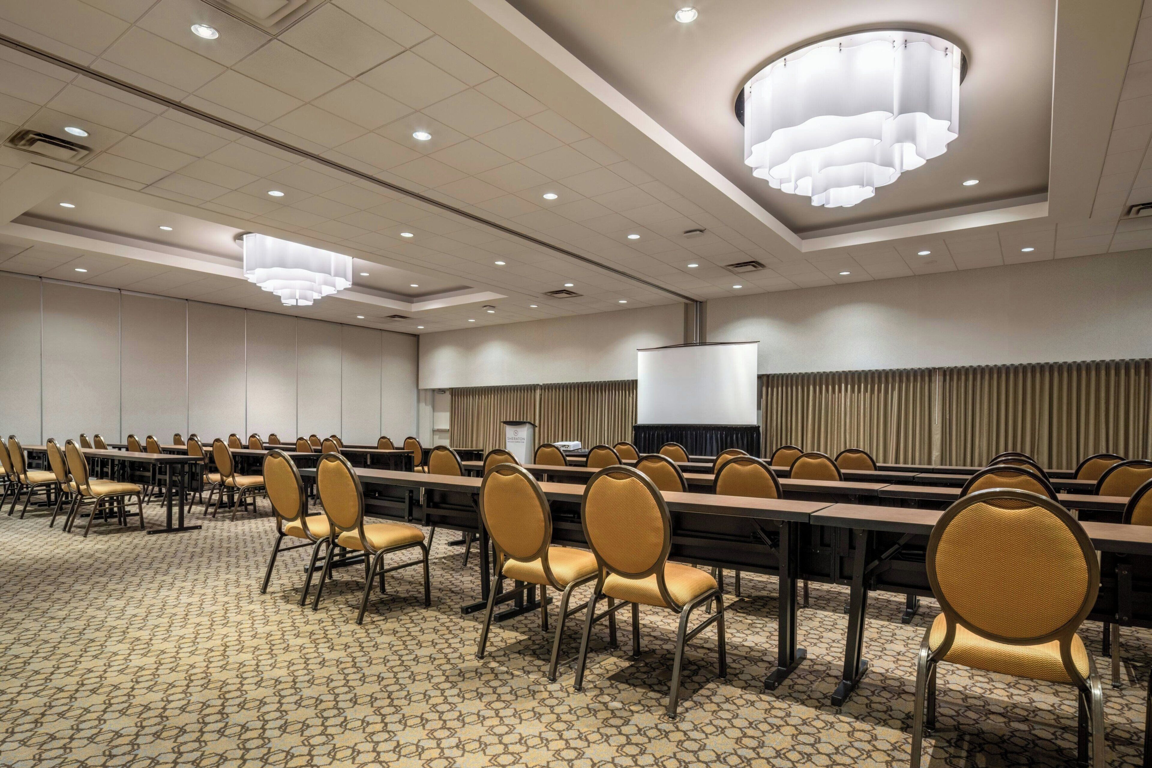 Un salón de conferencias con mesas largas, sillas y una pantalla de proyector.