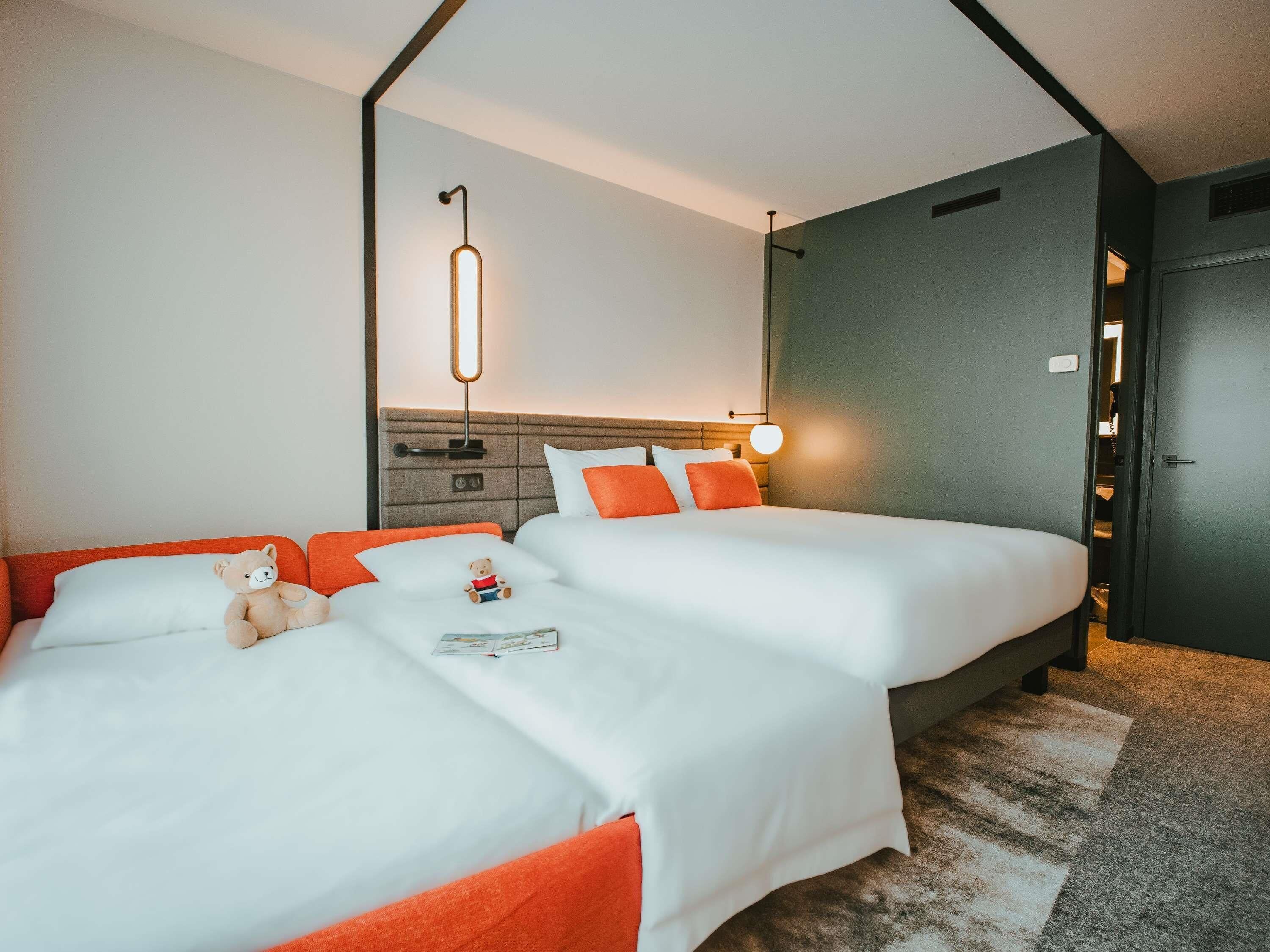 Une chambre d’hôtel avec deux lits, une table de chevet, une lampe fixée au mur et un ours en peluche sur l’un des lits.
