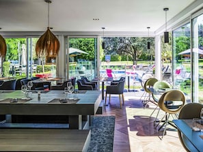 Un área de comedor al aire libre con mesas y sillas, ventanas amplias y vistas a una piscina y follaje.