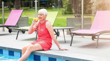 Una persona sentada junto a una piscina, con sillones de descanso y una mesa al fondo.