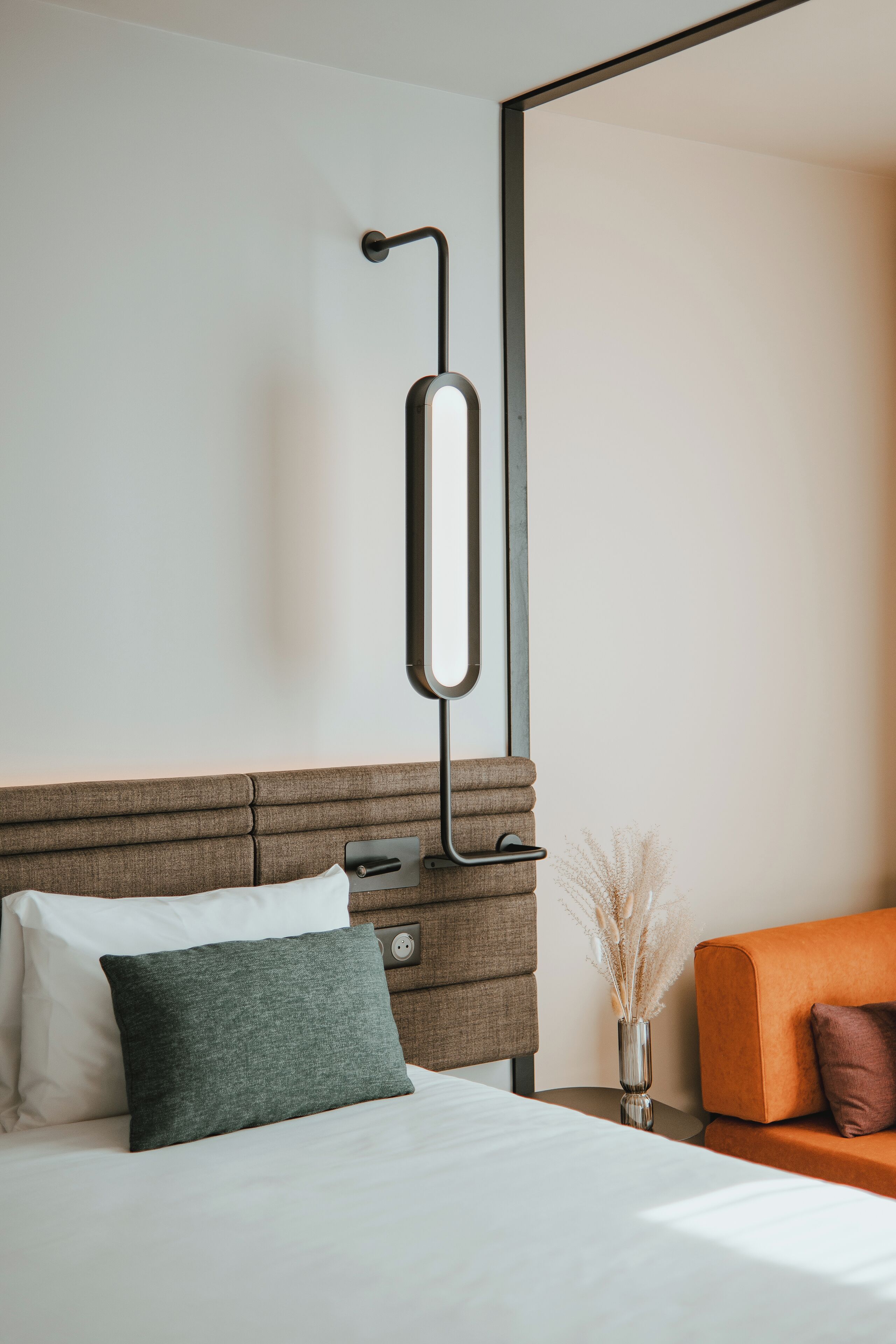 A modern hotel room with a bed, a nightstand, a vase with dried plants, and an orange armchair.