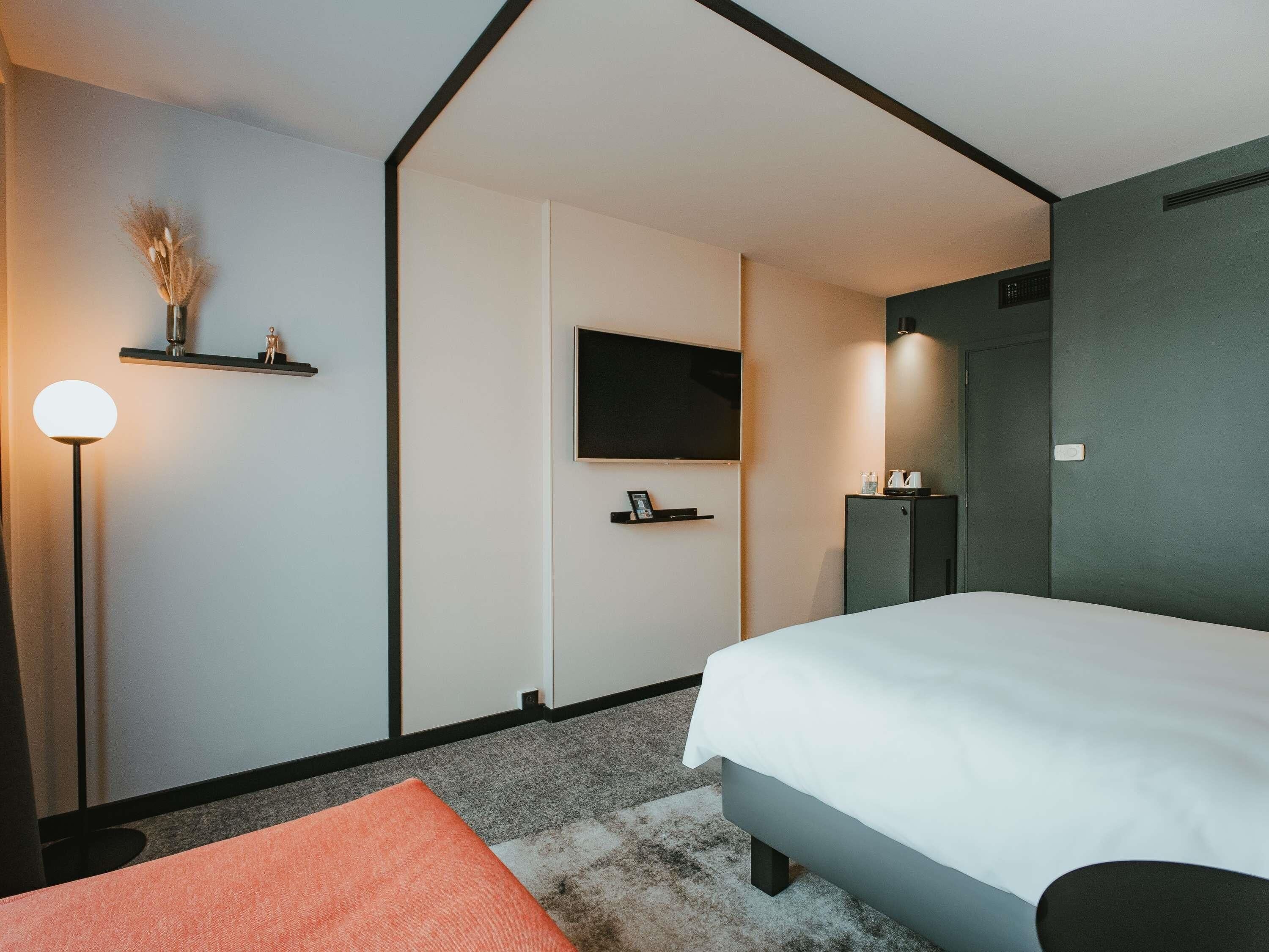 Een moderne hotelkamer met een bed, een flatscreen televisie, een nachtkastje, een lamp en een klein plankje met decoratieve objecten.