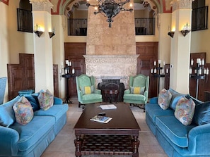 A spacious lobby with a high ceiling, ornate columns, and a fireplace. There are two green armchairs and a blue sofa with colorful cushions.