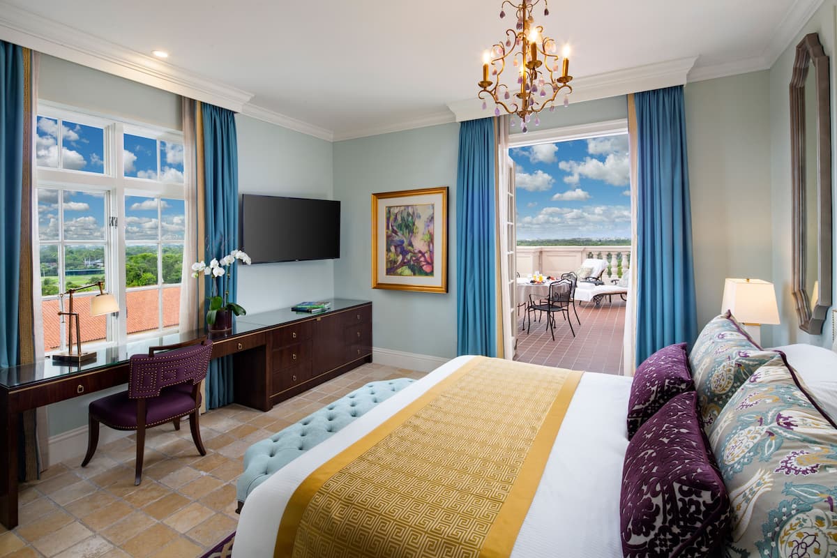 A hotel room with a large bed, a desk with a chair, a television, and a balcony with a view.