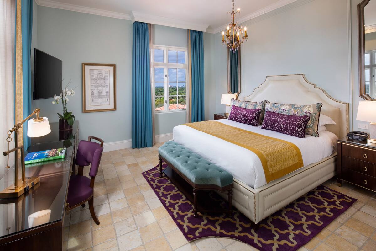 A bedroom with a large bed, a desk with a chair, a television, and a window with curtains.