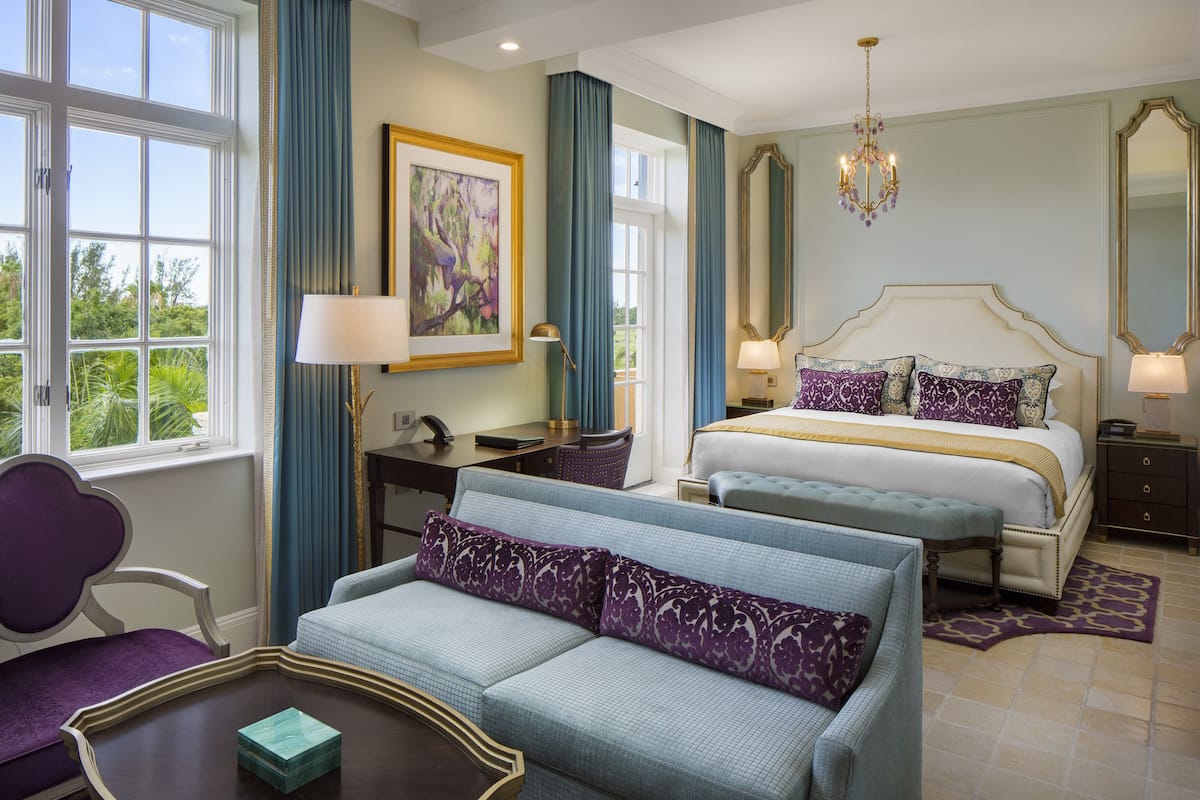 A hotel room with a large bed, a sofa, a desk, and a chair. There is a window with a view of greenery outside.
