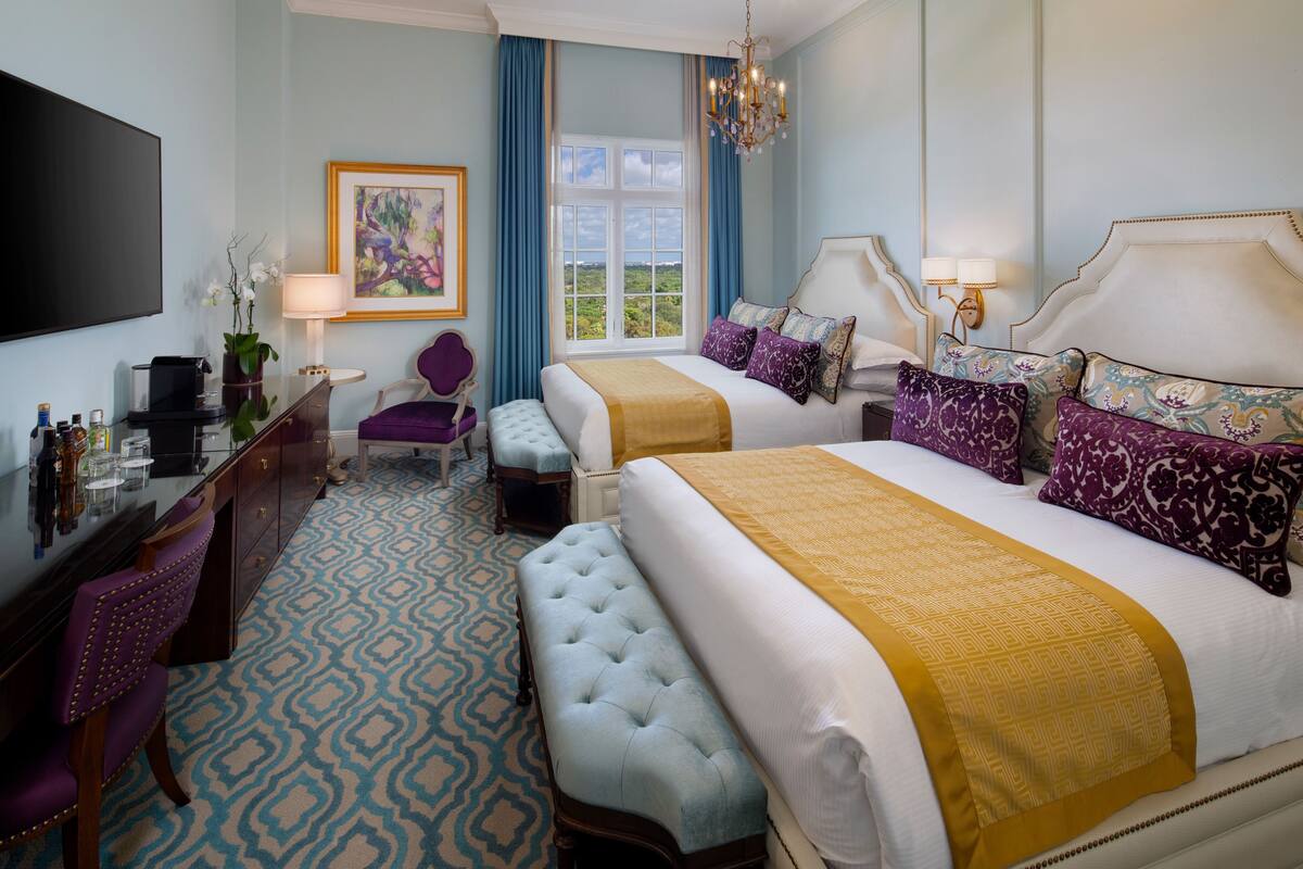 A hotel room with a large bed, a smaller bed, a desk with a chair, a TV, and a window with a view.