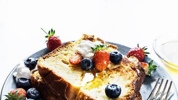 A slice of French toast topped with whipped cream, strawberries, and blueberries, served on a plate with a fork and a glass of a clear liquid.
