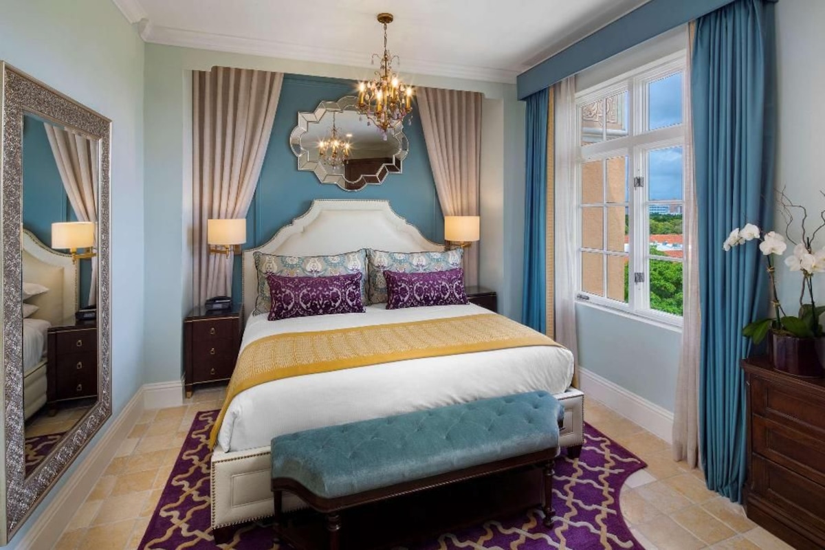 A bedroom with a large bed, two bedside tables, a chandelier, and a view of the outdoors through a window.