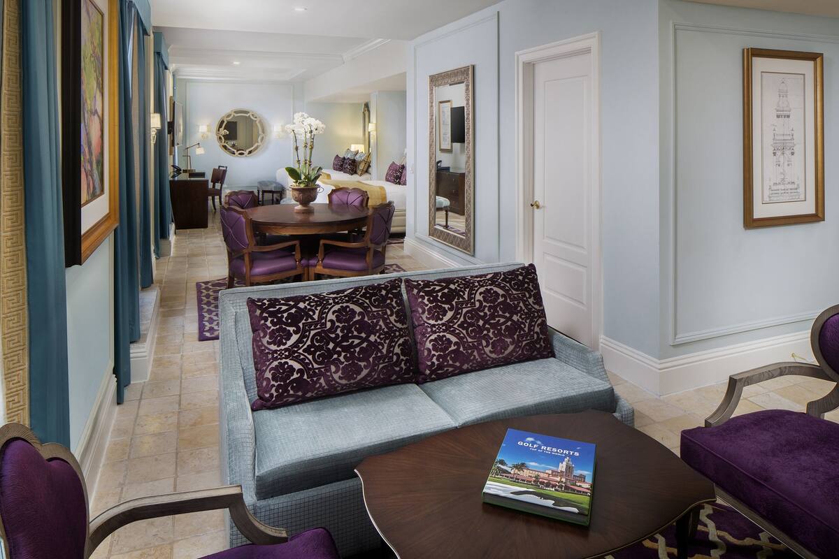 A hotel room with a sofa, a dining table with chairs, a coffee table with a book, and a large mirror on the wall.