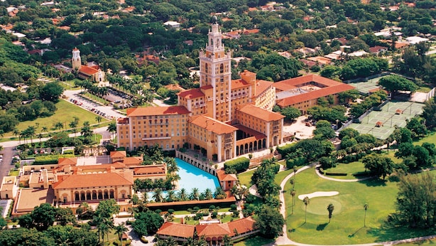 Vista aérea de un edificio grande e histórico con una torre destacada, un patio central con piscina y abundante vegetación circundante.