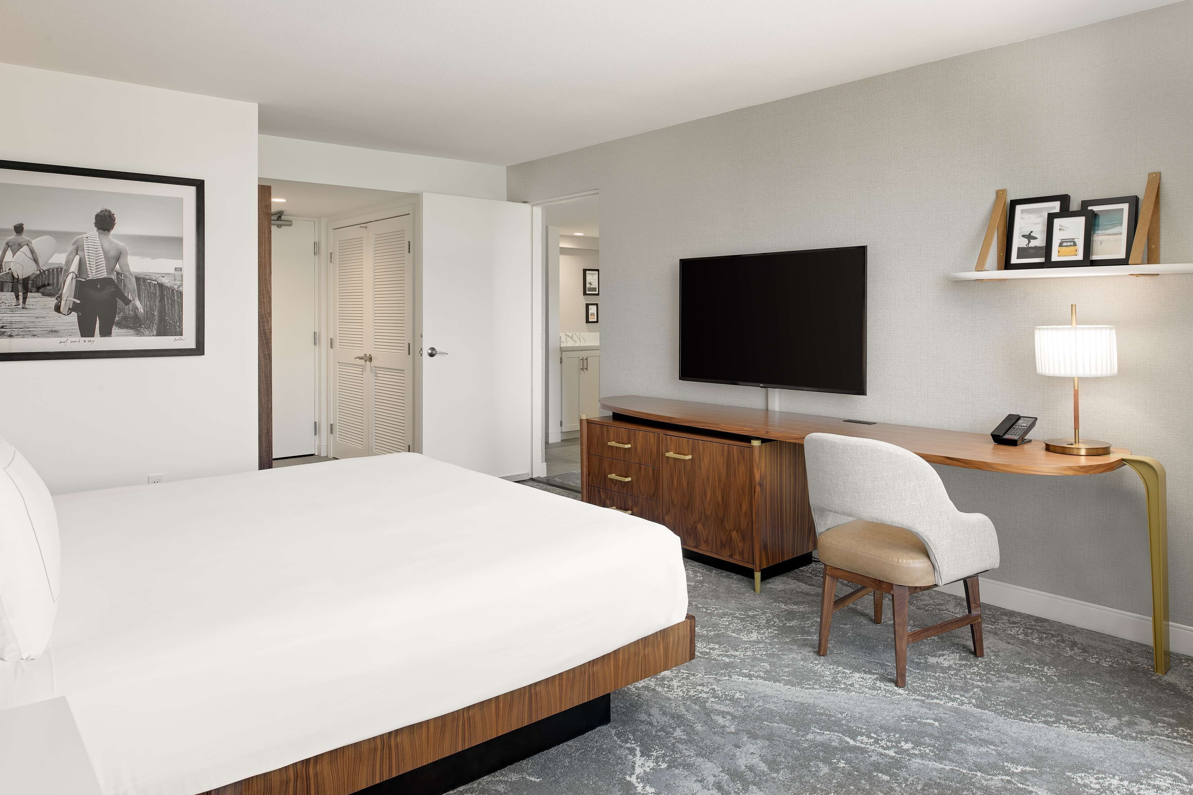 A hotel room with a bed, a desk, a chair, a TV, and a shelf with framed pictures.