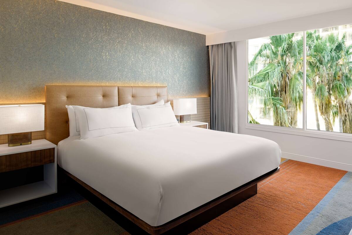 Une chambre d’hôtel avec un grand lit, deux tables de chevet, une commode, une fenêtre donnant sur des palmiers et un mur à motifs.